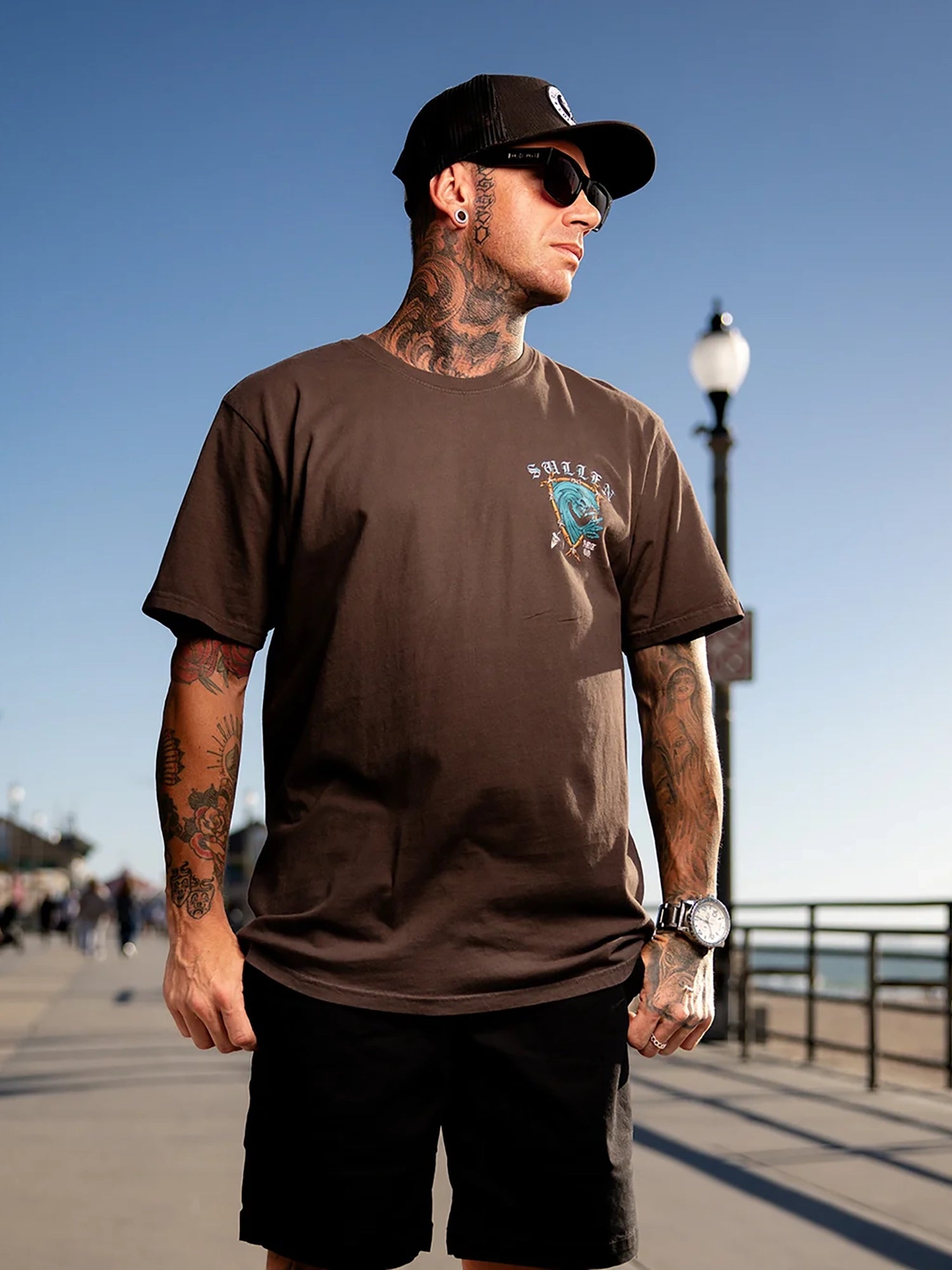 A man wearing a brown crewneck t-shirt with a graphic on the left chest, black shorts, a black baseball cap, and sunglasses, standing on an outdoor boardwalk.