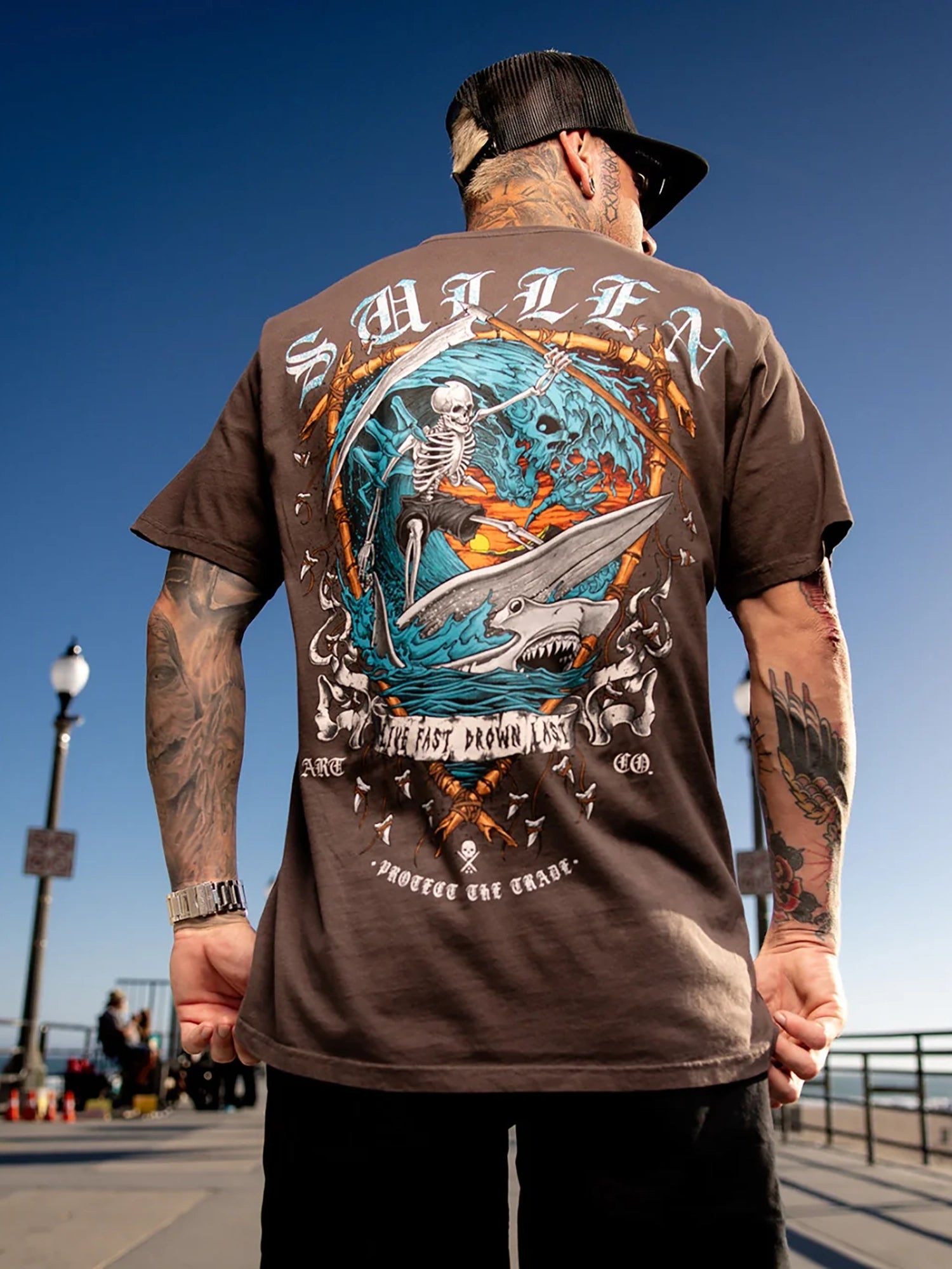 A man viewed from behind wearing a brown t-shirt with a large graphic print on the back. The graphic features a skeleton riding a surfboard on a blue ocean wave with the text 'SMILF' and 'LIFE IS BETTER WITHOUT GRAPE'.