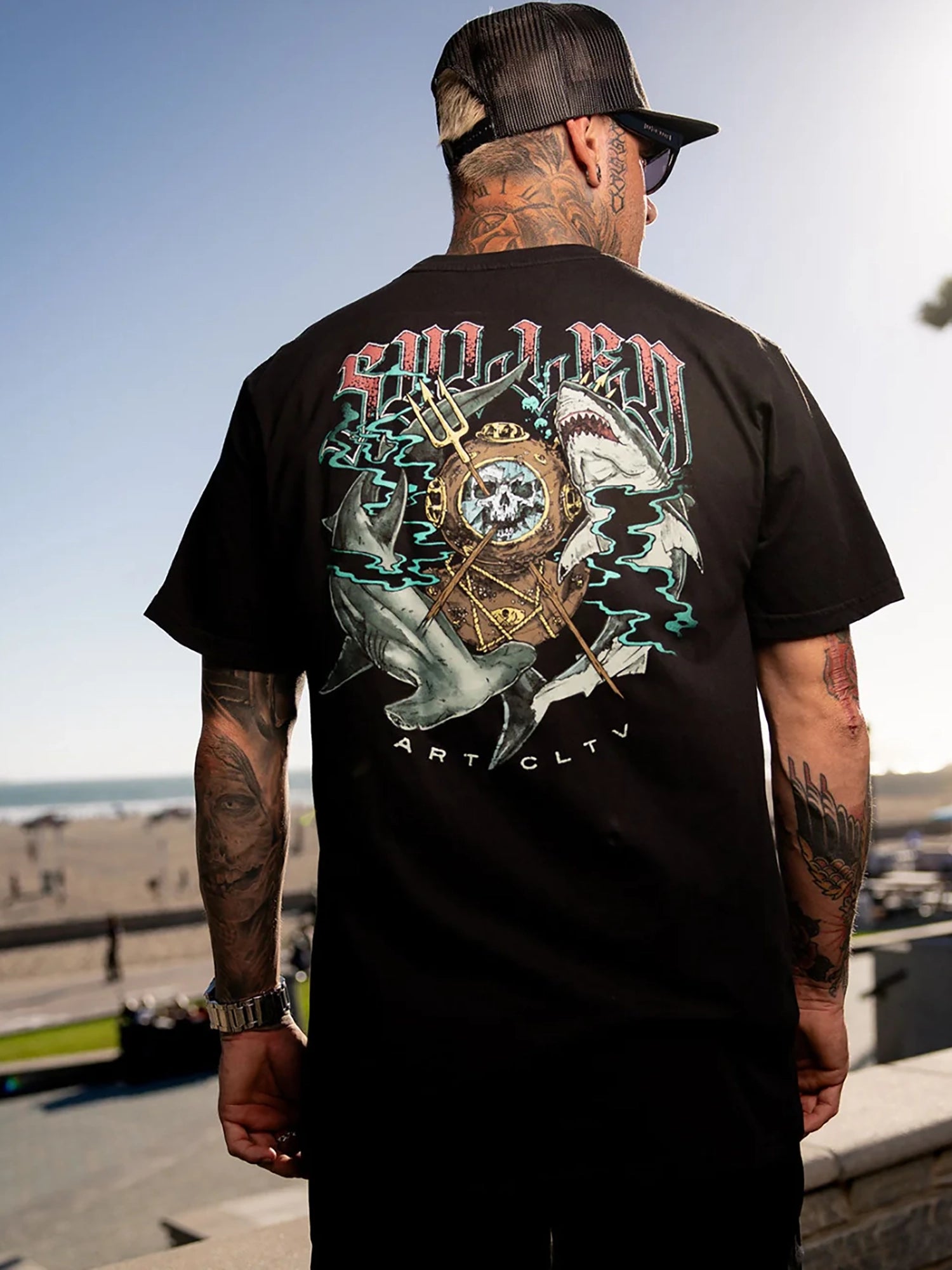 A man viewed from behind wearing a black t-shirt with a large graphic print on the back, featuring sharks, dolphins, a skull, and the text 'SOLJEN ART CUTL'.