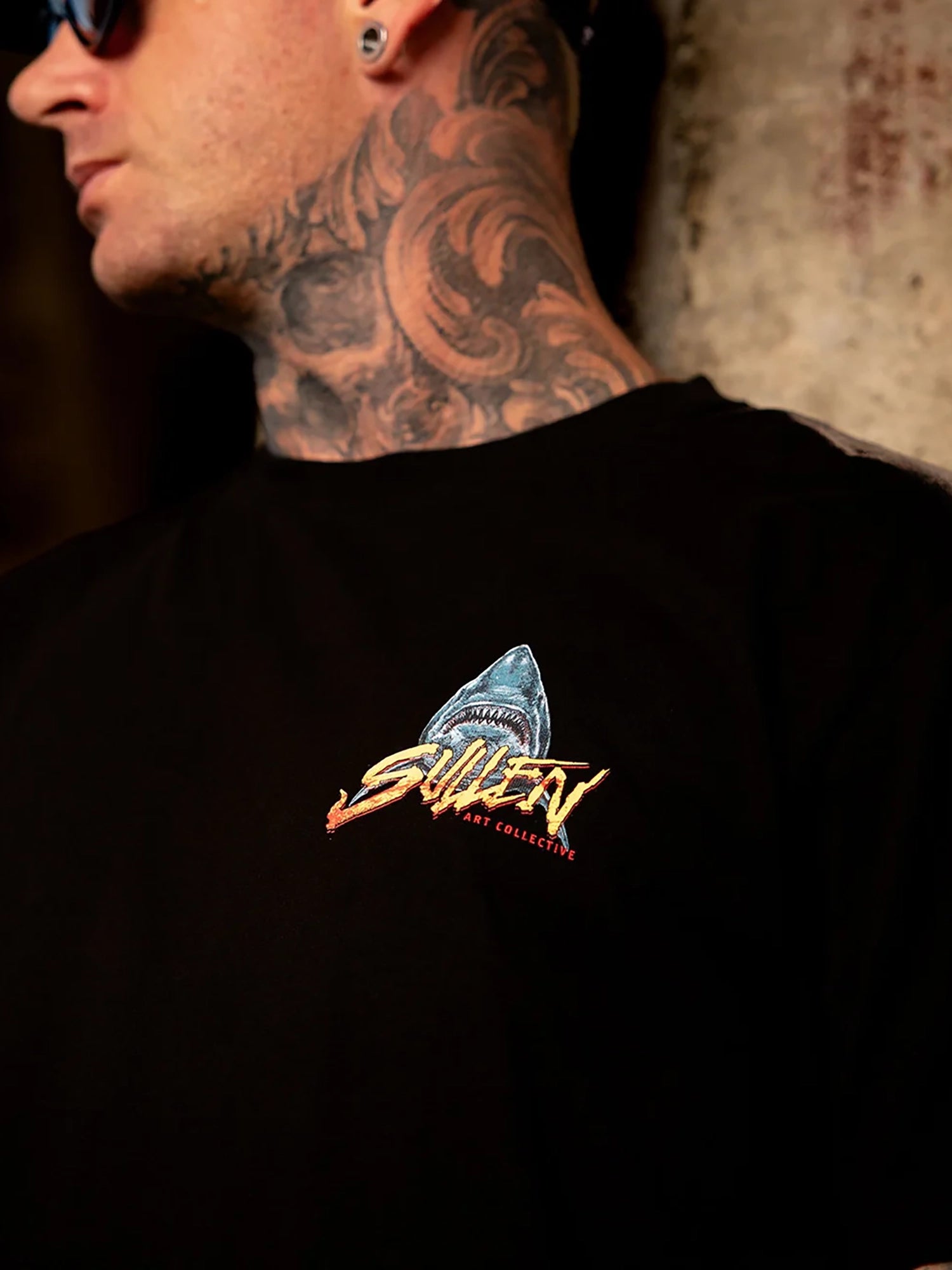 Close-up of a man wearing a black t-shirt with a colorful graphic on the chest. The graphic features a shark head with the word 'SURF' in stylized yellow and orange lettering, and 'SAN DIEGO' underneath.