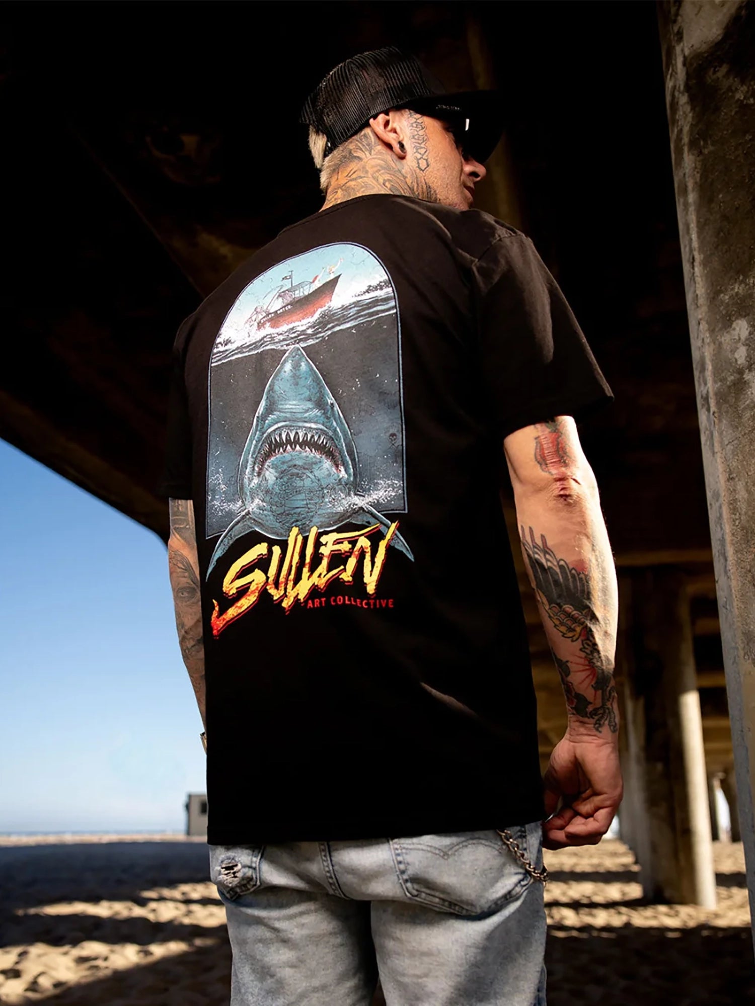 A man wearing a black t-shirt with a large graphic print of a shark and a ship on the back, standing under a concrete pier structure.