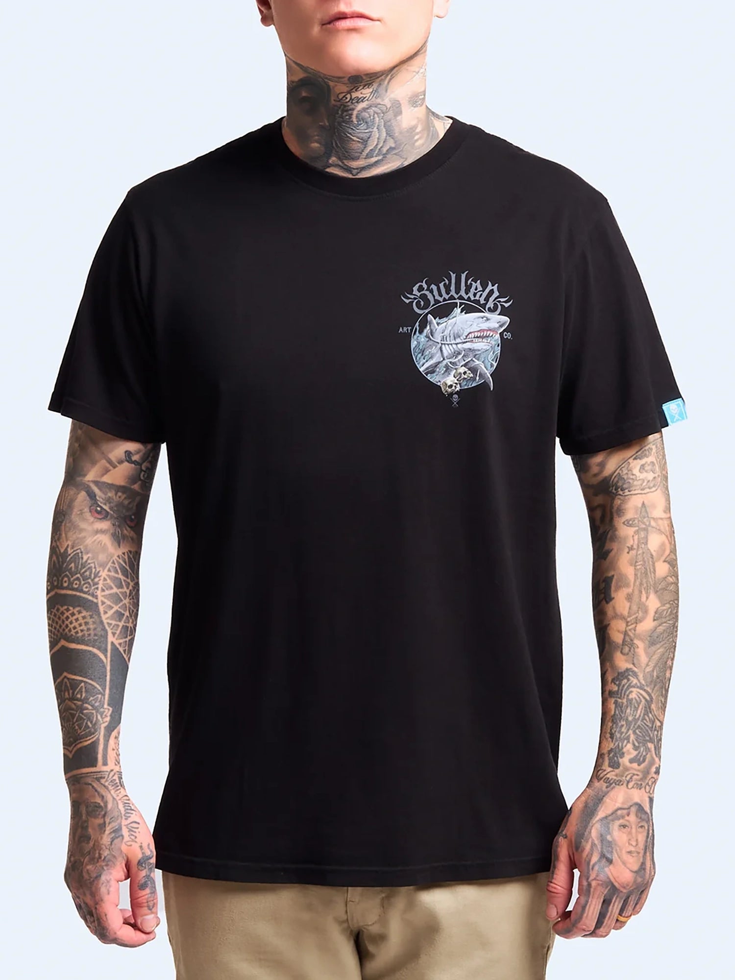 A man wearing a black short-sleeve t-shirt with a white and gray graphic on the left chest that reads 'Gullion'. The model has visible tattoos on his neck, arms, and hands, and is wearing khaki pants against a light blue background.