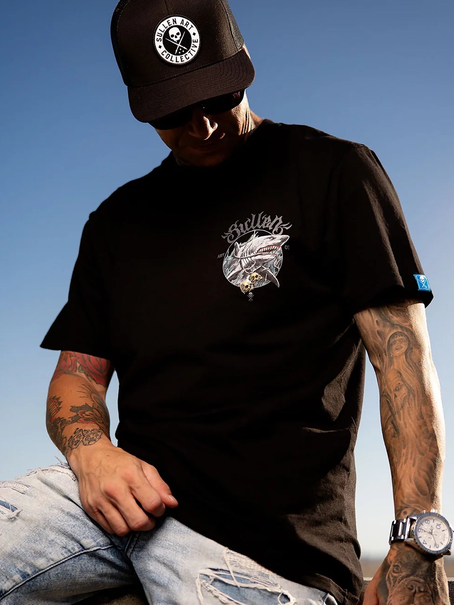 A man wearing a black t-shirt with a graphic print on the chest, a black cap with a circular logo, and light-wash ripped jeans, photographed against a clear blue sky.
