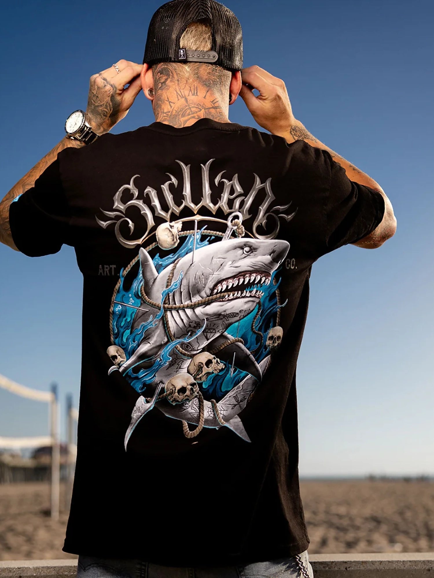 A man viewed from behind wearing a black t-shirt with a large graphic print of a shark skeleton and the word 'Sicilian' in stylized white lettering. The background shows a sandy beach and blue sky.