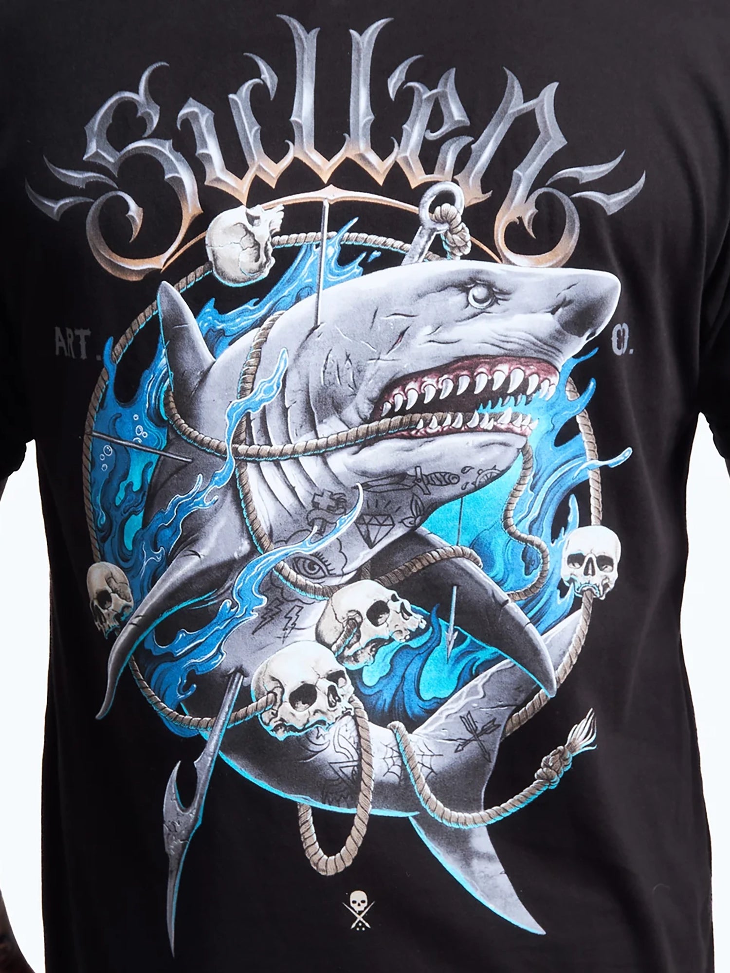 A close-up of a black graphic t-shirt featuring a large design of a shark wrapped in ropes with skulls and blue flame-like accents, under a stylized metallic 'Sullen' logo.