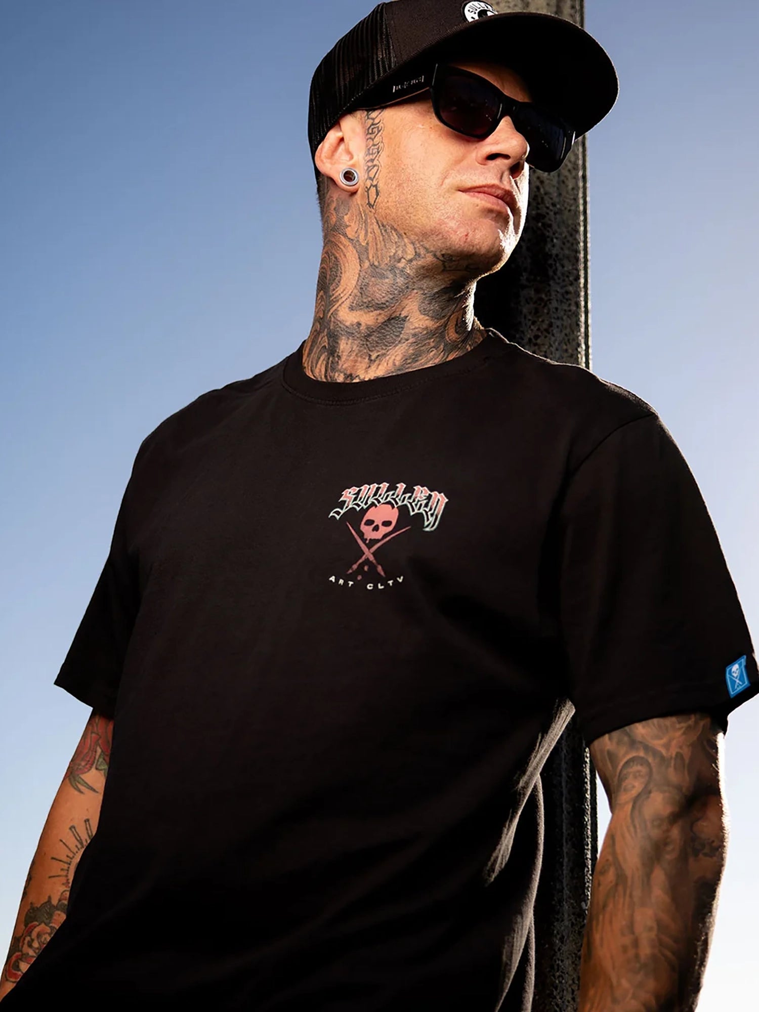 A tattooed man wearing a black crew-neck t-shirt with a skull and crossbones graphic on the chest, a black baseball cap, and sunglasses, posing against a clear blue sky.