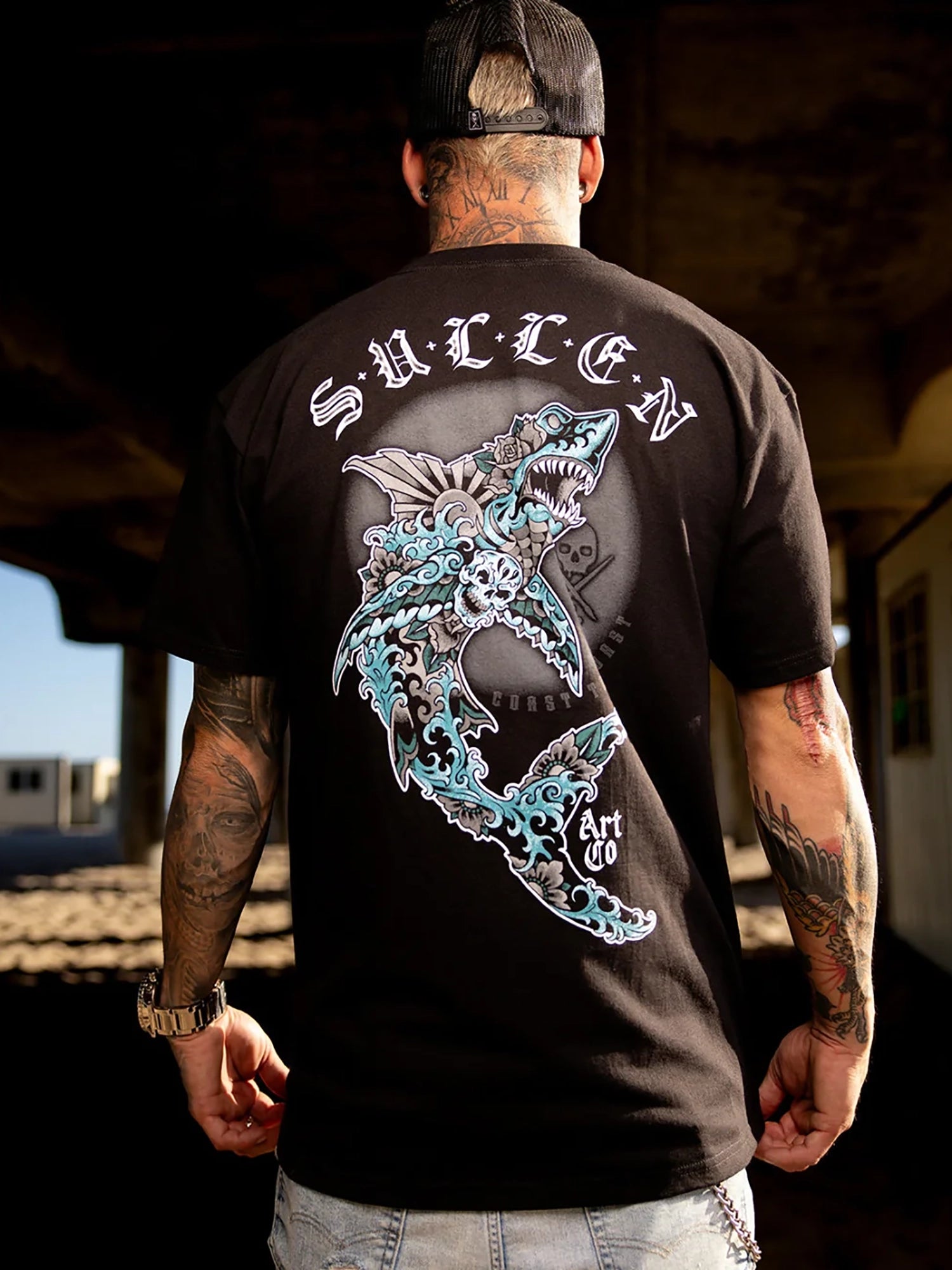 A man viewed from behind wearing a black t-shirt with a large graphic of a shark and the word 'SURF' on the back, standing in a dim outdoor setting.