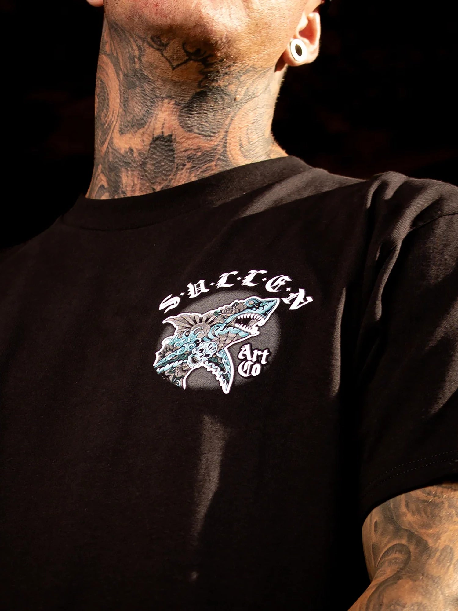 A close-up of a person wearing a black t-shirt with a graphic on the chest. The graphic features the word 'SUFFER' in a stylized white font above a detailed illustration of a shark in shades of blue, white, and gray.