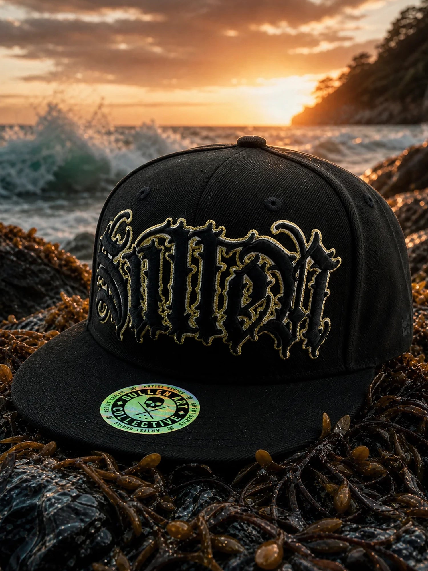 A black flat-brim cap with ornate gold embroidered lettering on the front, resting on wet rocks covered in seaweed at sunset by the ocean. A circular green and white sticker is visible on the brim.